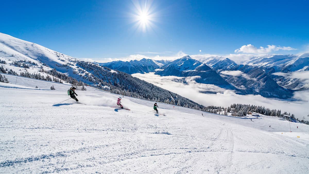 Drie skiërs trekken krachtige bochten door de poedersneeuw op de zonnige pistes van de Wildkogel-Arena, met een adembenemend uitzicht op de besneeuwde toppen van de Hohe Tauern – wintersportplezier in de perfecte wintervakantie.